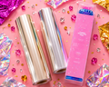 Two rolls of metallic foil and a box of 'Ugly Betty' brand foil on a pink background with decorative elements.