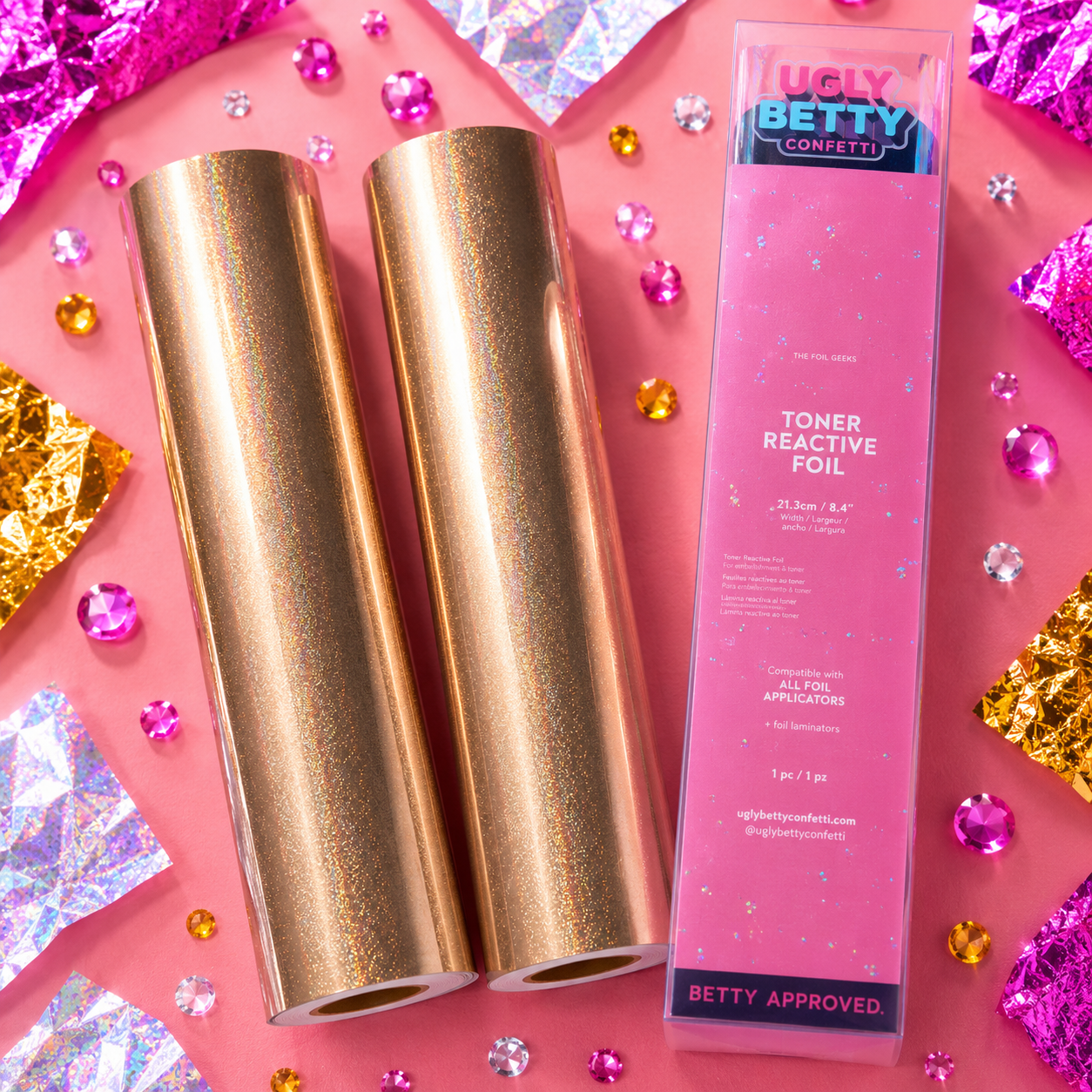 Gold foils and a pink packaging tube labeled 'Ugly Betty Confetti' on a pink background with confetti.