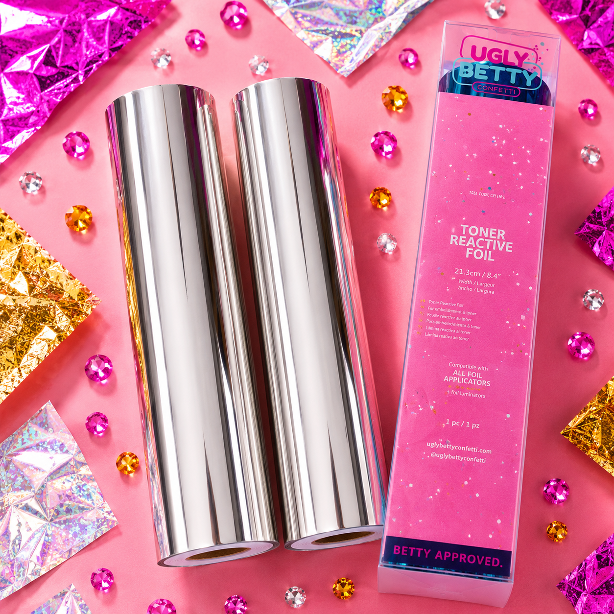 Two rolls of silver foil and a box of Ugly Betty toner reactive foil on a pink background with decorative crystals.