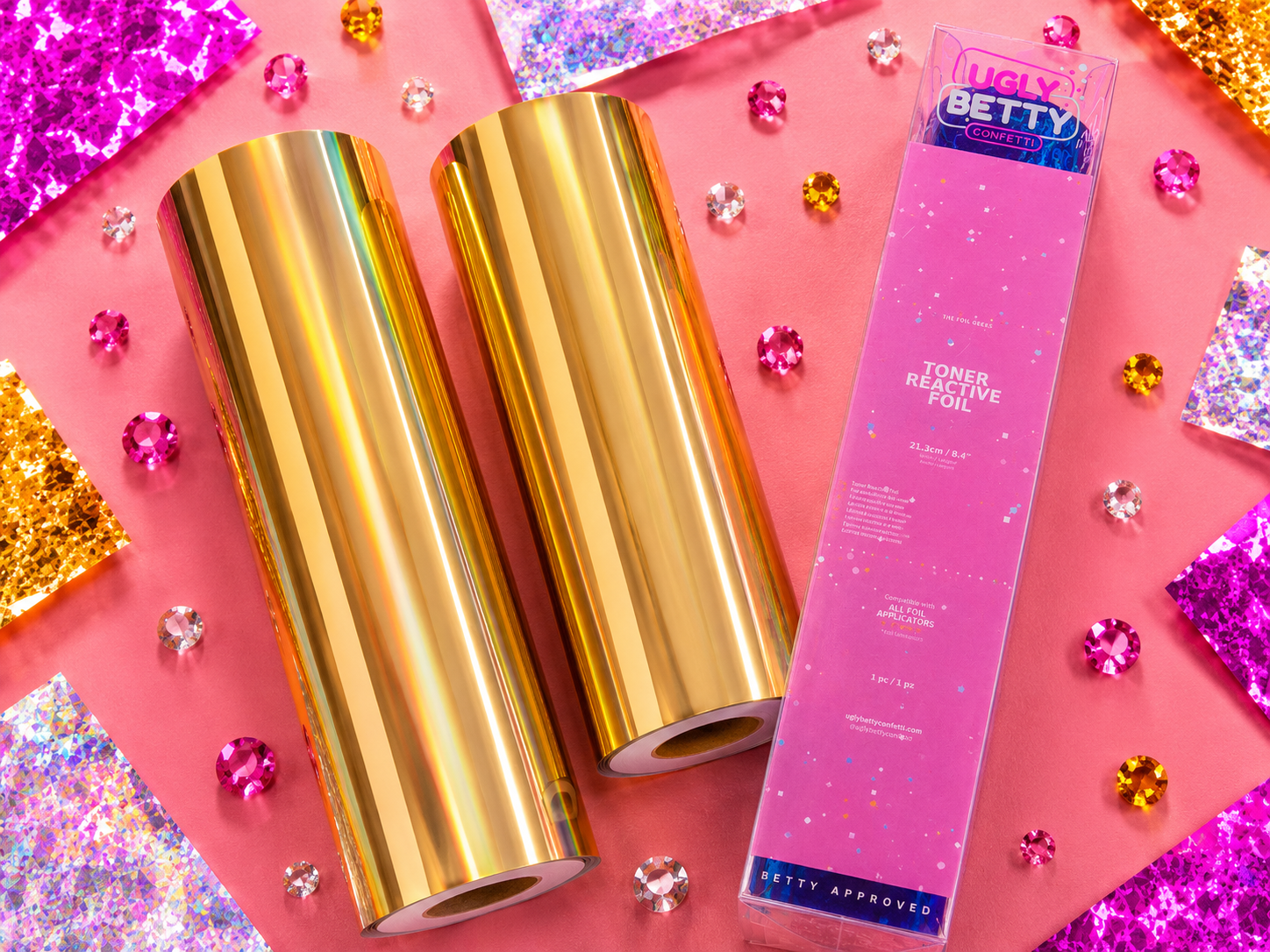 Gold foil rolls and a pink packaging box labeled 'Ugly Betty' on a colorful background with glitter.