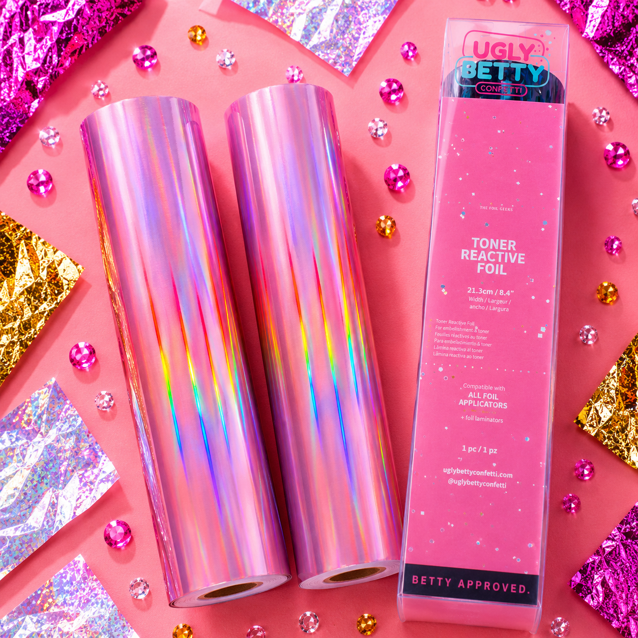 Two rolls of iridescent foil and a box labeled 'Ugly Betty' on a pink background with decorative elements.