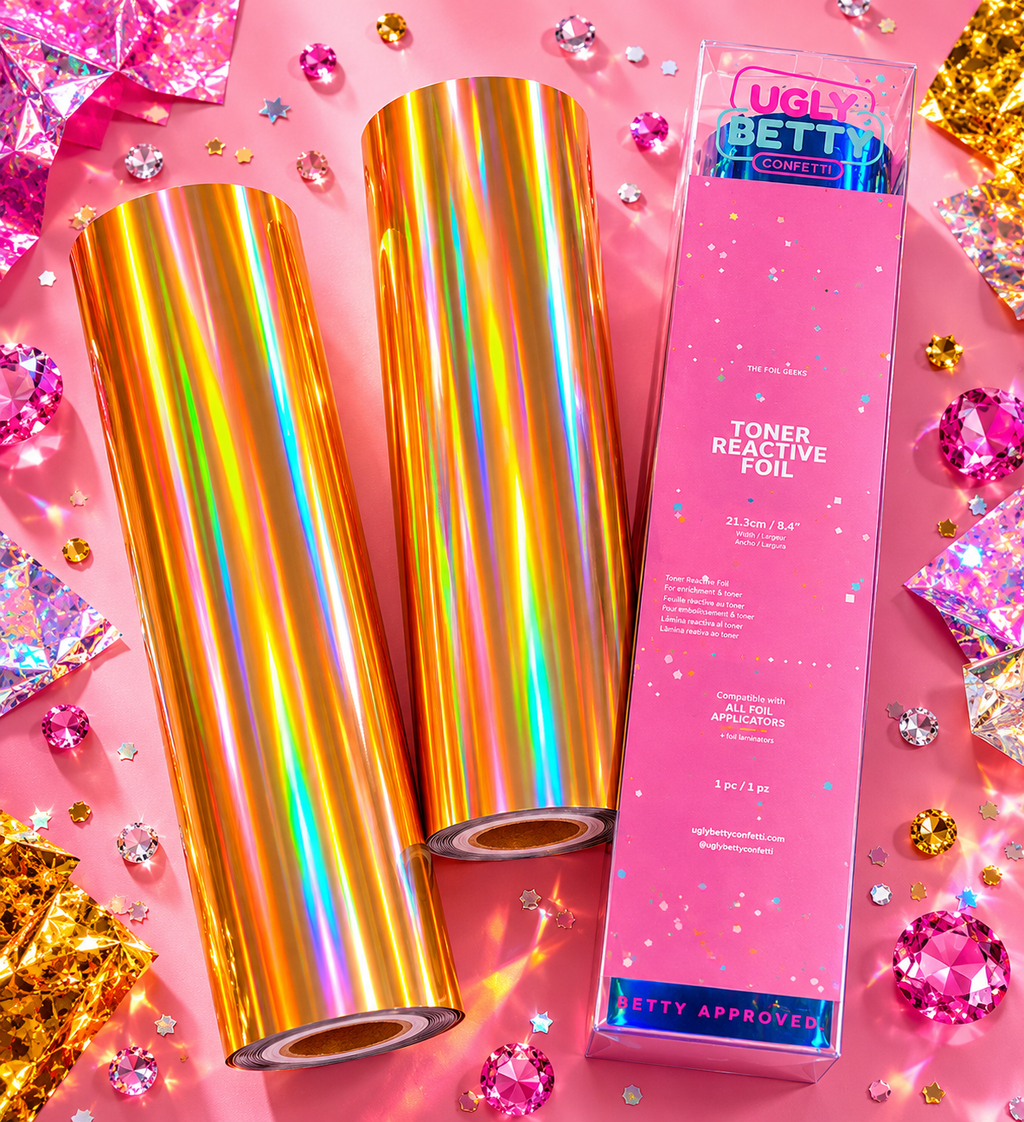 Two rolls of colorful toner reactive foil with a pink packaging on a pink background with confetti and gemstones.